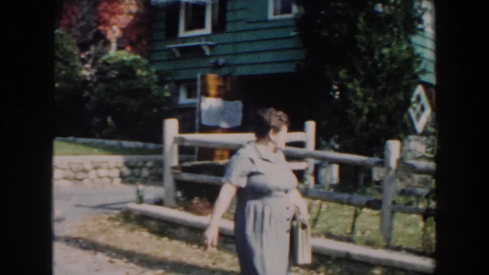 In stated 1960 Miami footage, a woman walks along a fence in front of a green house and landscaped yard.