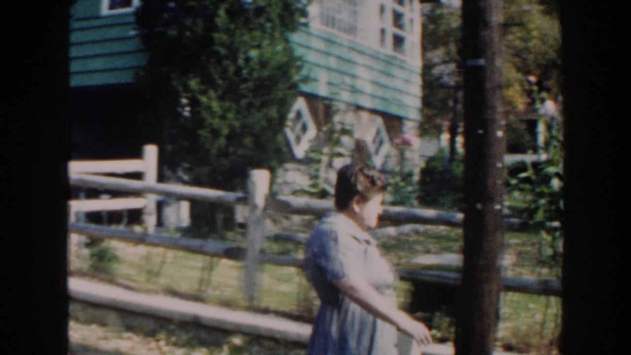 Woman passes a tree and fence with the green house and garden behind her.