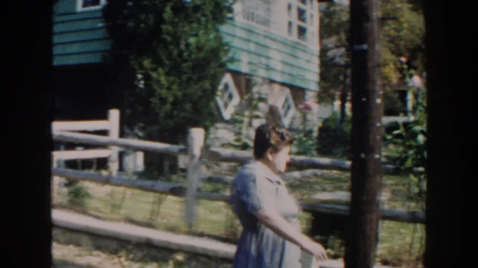 The woman continues walking past a tree and split-rail fence in the Miami garden scene.
