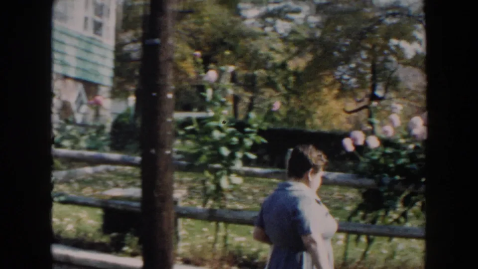 The woman turns away from the camera near the fence, with lawn, trees, and pink flowers visible in the yard.