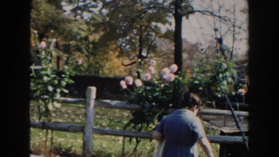 Near the end of the clip, the woman walks away through the garden area beside a fence and pink flowering shrubs.