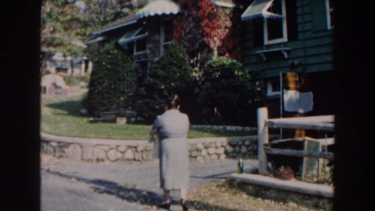 Woman stands near a driveway, stone wall, and green house exterior.