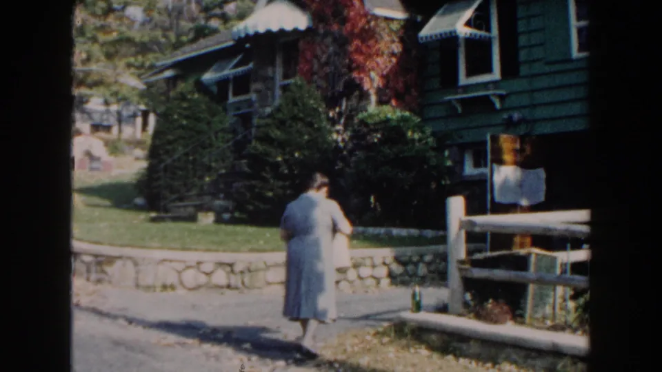 The woman in a blue dress walks across the yard near a stone wall, driveway, shrubs, and the exterior of a green house.
