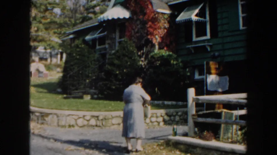 In the final frame, the woman bends or reaches near the grass with the green house, stone wall, and wooden railing behind her.
