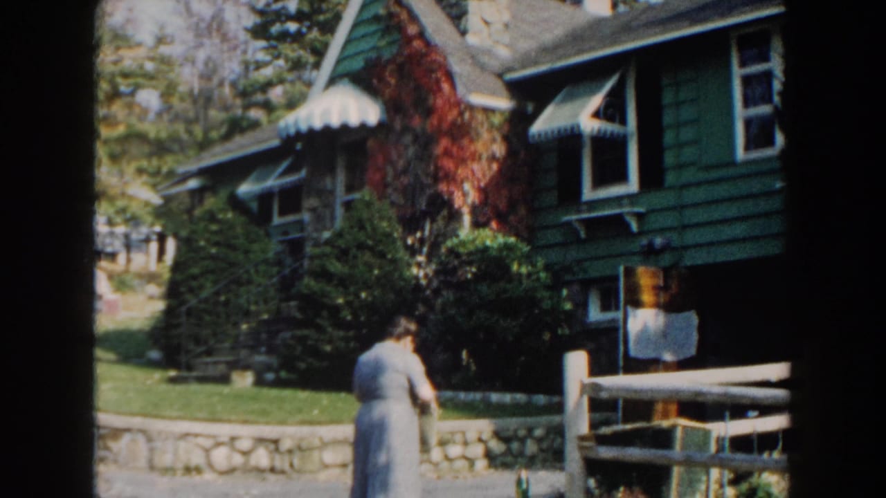 1960 Miami home-movie frame showing a woman approaching a green house with white trim, awnings, ivy, lawn, and fence.