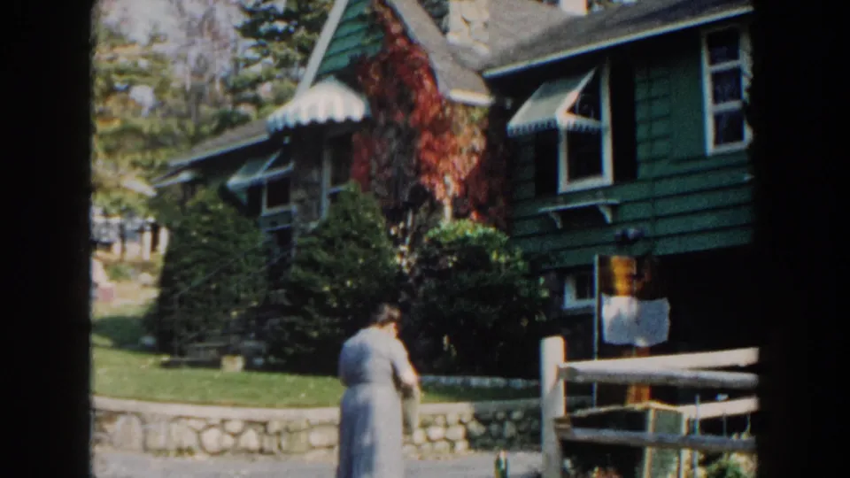 1960 Miami home-movie frame showing a woman approaching a green house with white trim, awnings, ivy, lawn, and fence.