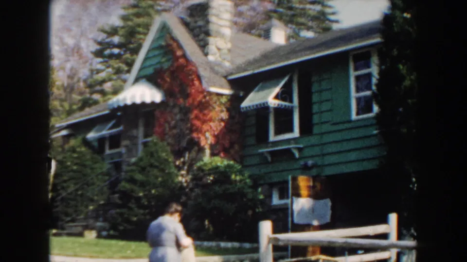1960 Miami exterior view of a green residential house with striped awnings, red ivy, trees, lawn, fence, and a woman near the front.