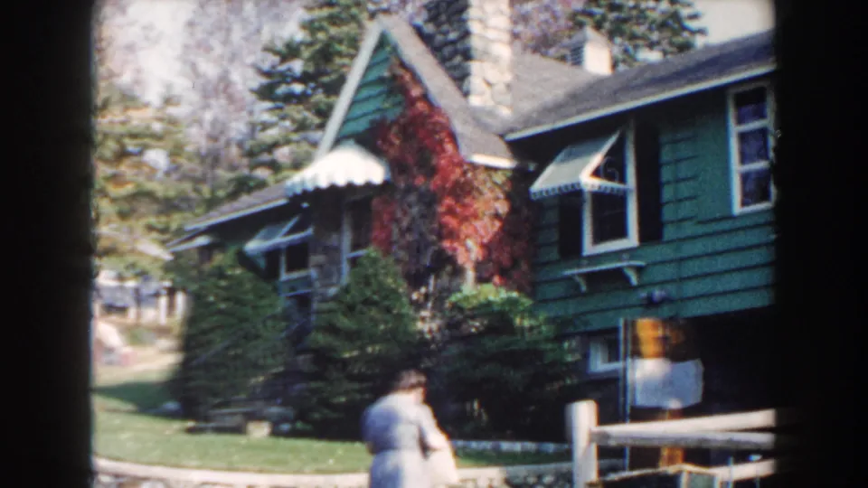 1960 Miami home-movie view of the green house exterior, showing awnings, red ivy, a chimney, lawn, trees, and a woman near the foreground.