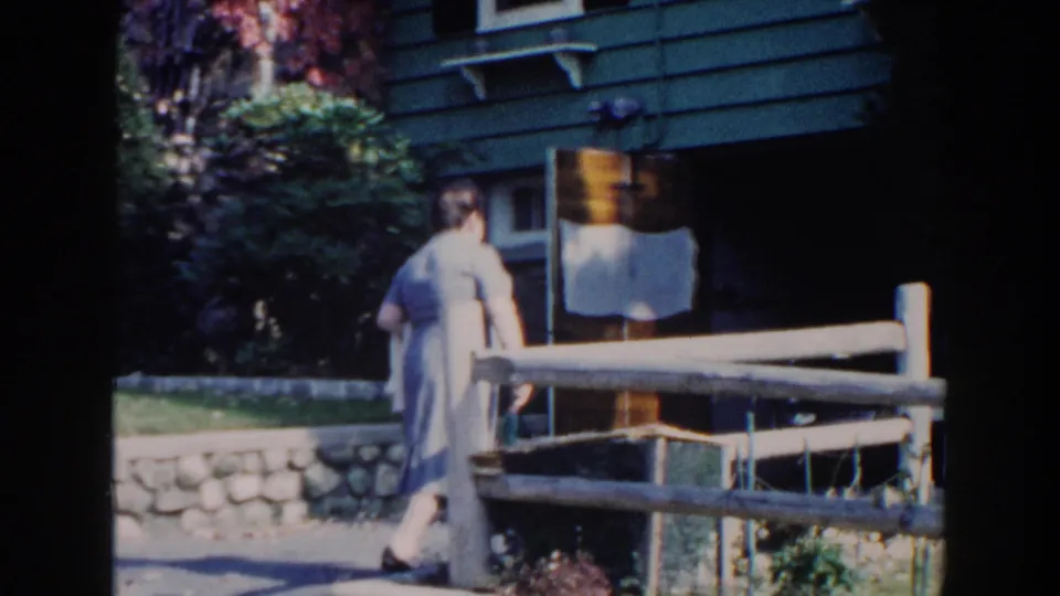 1960 Miami frame showing a woman from behind near a wooden fence, with the green house, white awnings, red ivy, shrubs, and lawn behind her.
