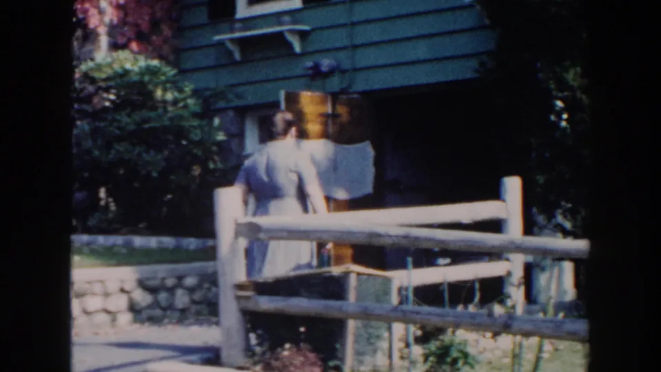 In 1960 Miami home-movie footage, a woman stands outside a green-sided house behind a rustic wooden fence.