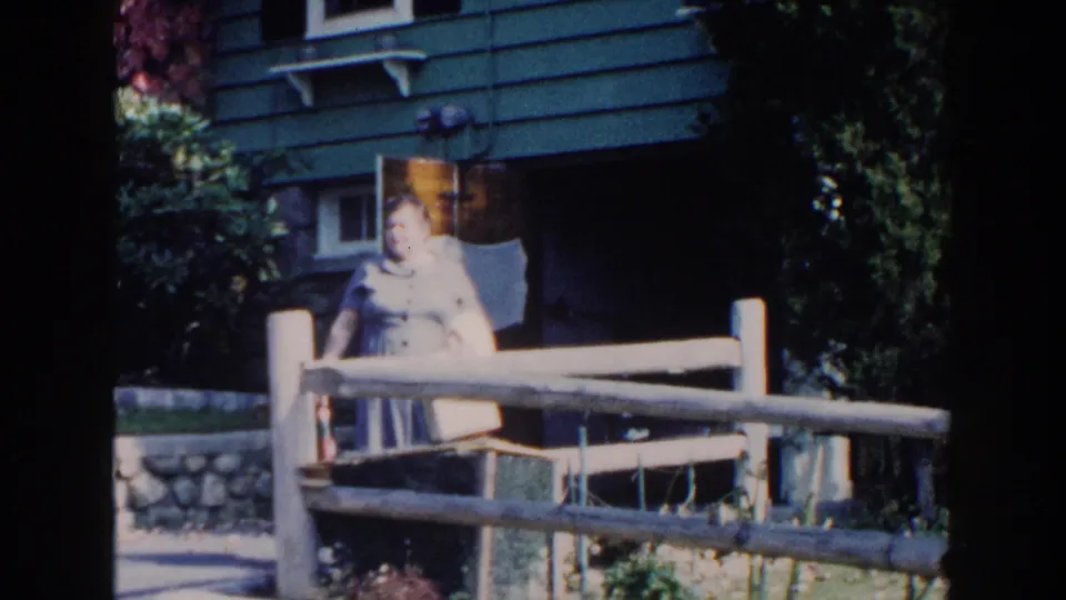 The woman remains near the fence in the sunlit residential yard, with green siding, stonework, and shrubs behind her.