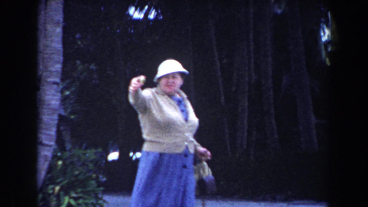 In 1960 Miami home-movie footage, an adult woman in a white hat, beige cardigan, and blue skirt gestures near a tree-lined walkway.