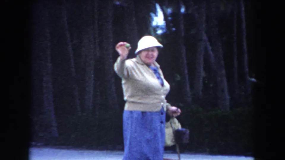 Adult woman in a white hat and blue skirt raises her hand while carrying bags in a shaded Miami outdoor setting.