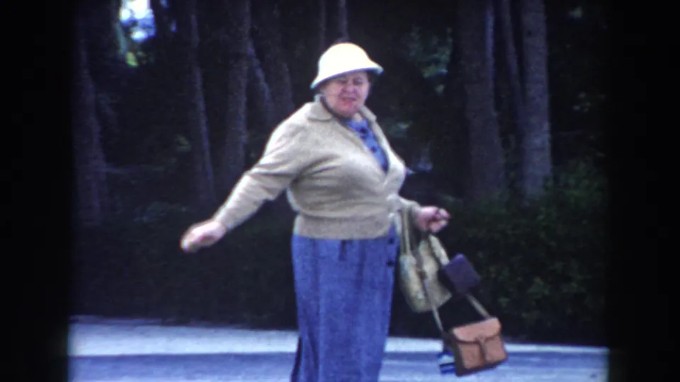 Adult woman in 1960 Miami walks along a pale path, wearing a white hat and blue skirt while holding small bags.