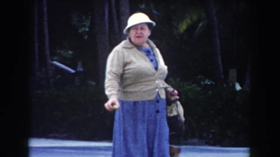 Adult woman in a white hat, beige cardigan, and blue skirt faces the camera while carrying bags in a shaded Miami street or park setting.