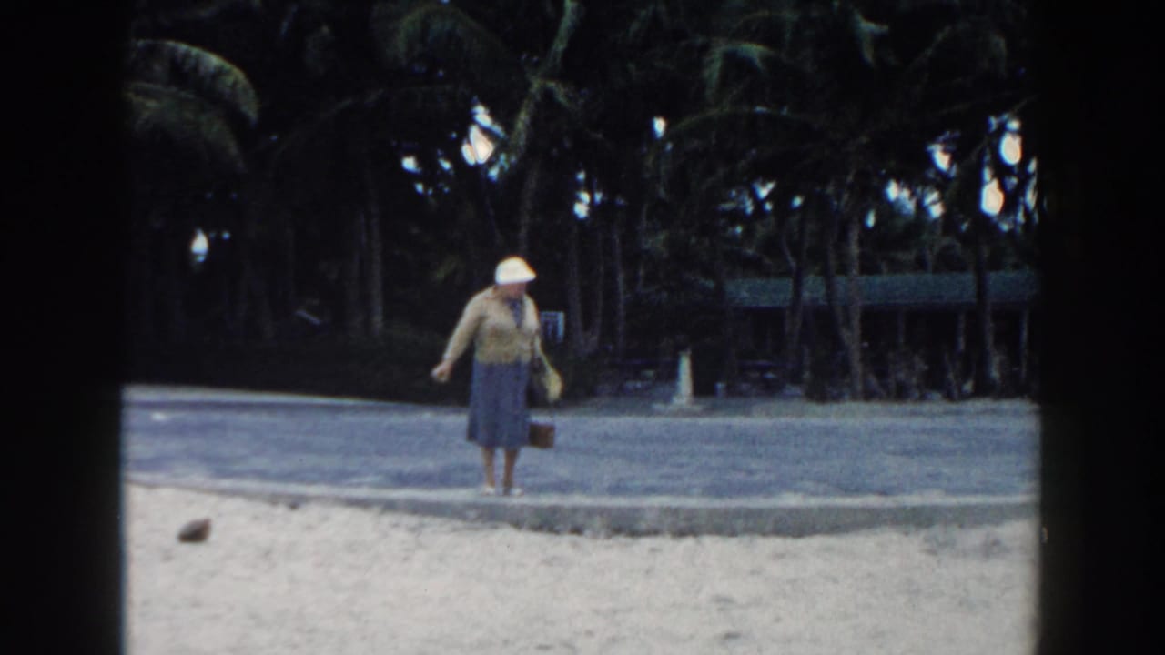 In 1960 Miami home-movie footage, a woman in a white hat and blue dress walks beside sand, a paved path, and palms.