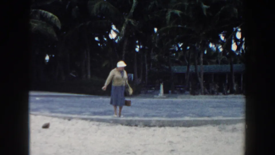 In 1960 Miami home-movie footage, a woman in a white hat and blue dress walks beside sand, a paved path, and palms.