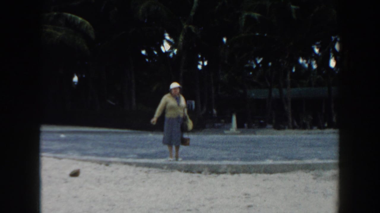 Woman stands near the beach path with palms behind her, holding a small bag.
