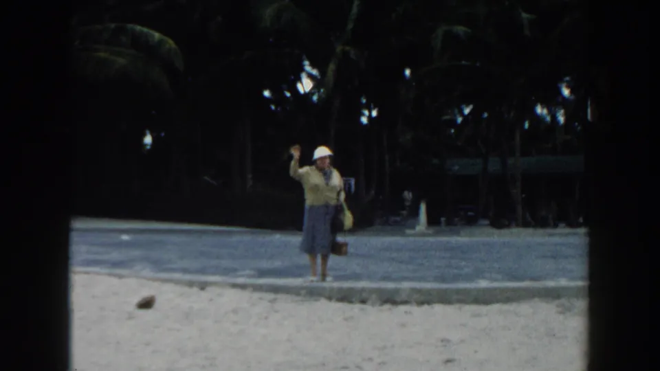 A woman in a white hat and blue dress waves toward the camera in 1960 Miami beach home-movie footage.