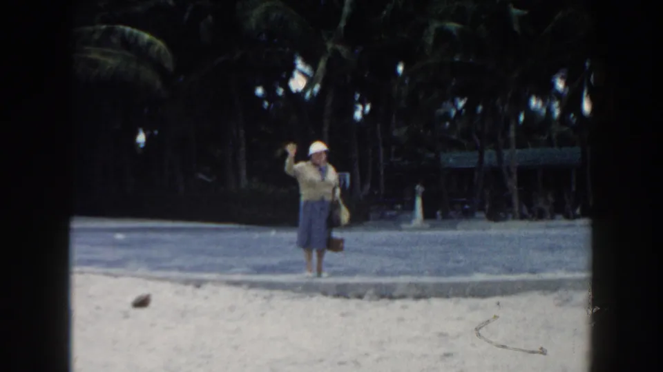 The final frame shows the woman still waving toward the camera, framed by sand, a paved path, and palms in Miami.