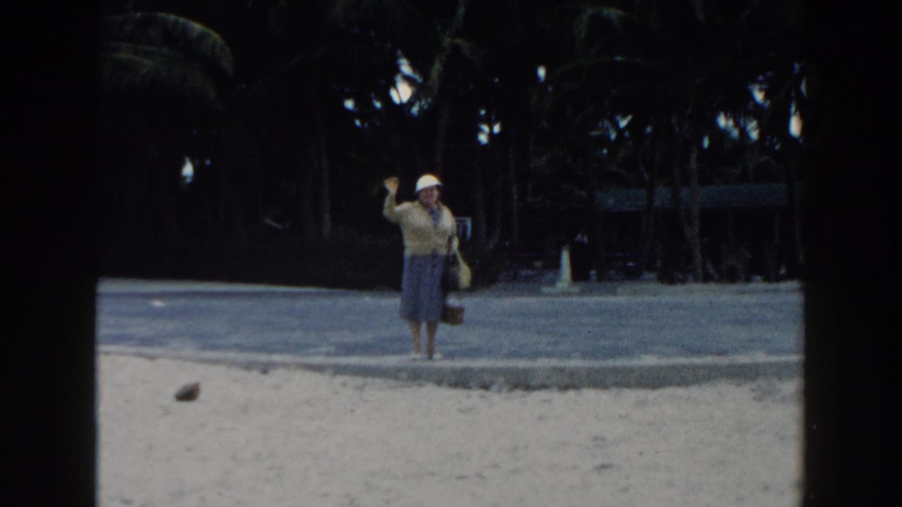 Older woman in a white hat waves while standing on a sandy beach near palm trees.