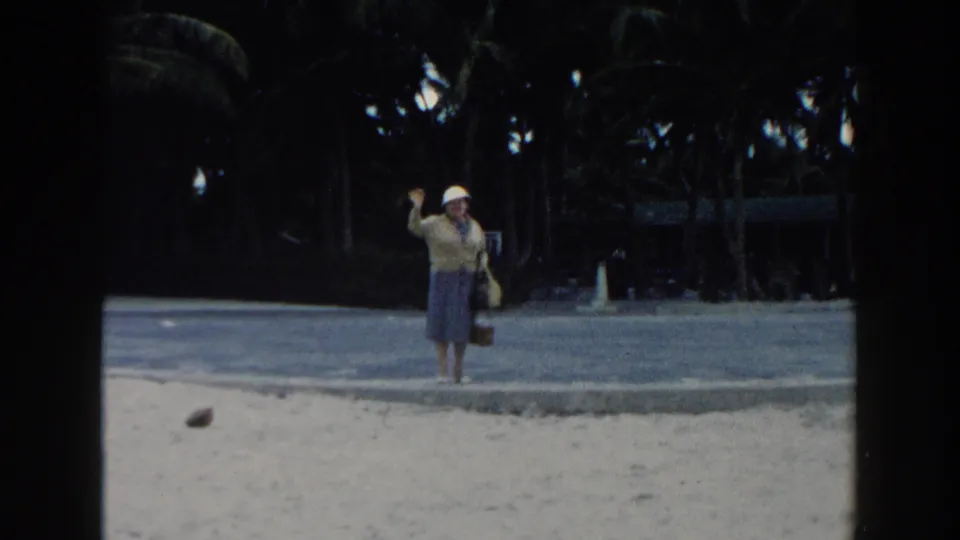 In 1960 Miami home-movie footage, an older woman in a white hat and skirt waves from a sandy beach beside a palm-lined path.