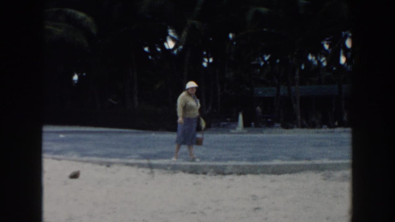 The woman walks along the sandy Miami beach in a white hat and blue skirt, with dark palm trees behind the paved edge.