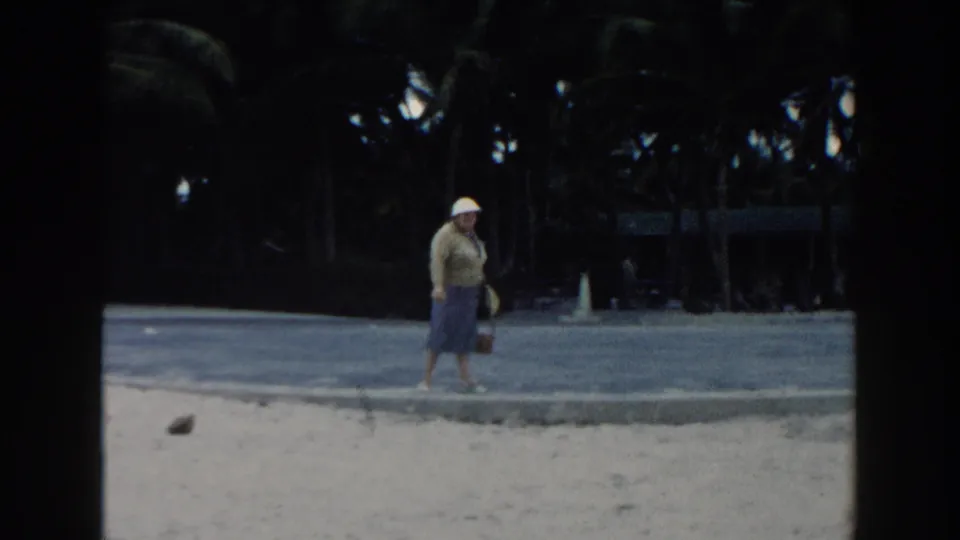 The woman walks along the sandy Miami beach in a white hat and blue skirt, with dark palm trees behind the paved edge.