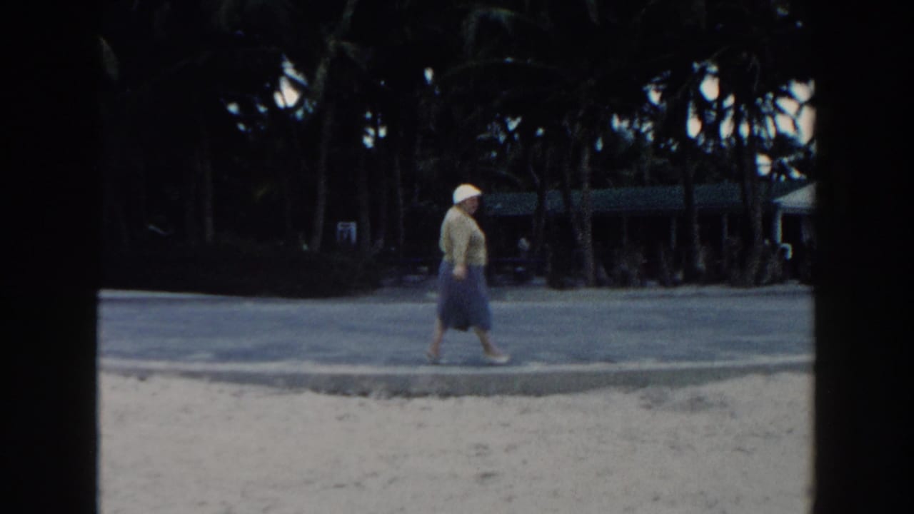 The 1960 Miami beach view shows the older woman walking past the camera's line of sight with palms and a shaded path behind her.