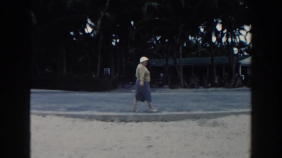 The 1960 Miami beach view shows the older woman walking past the camera's line of sight with palms and a shaded path behind her.
