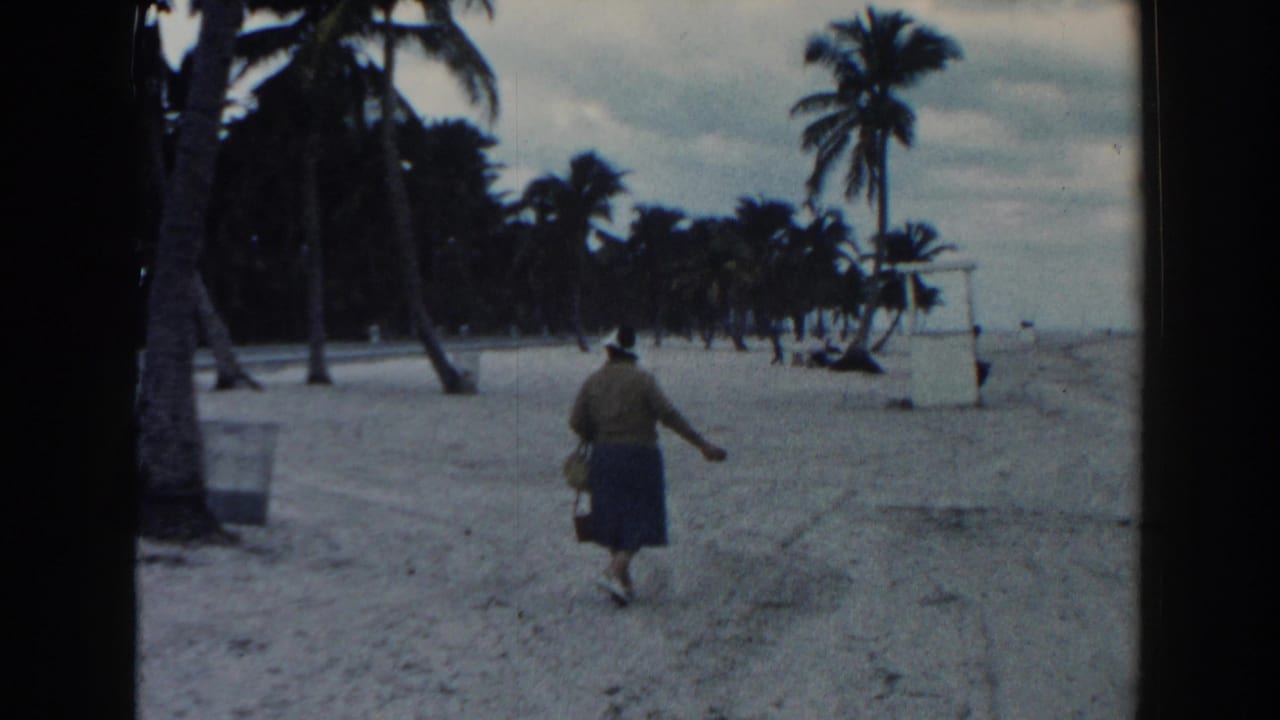 The woman walks along the Miami beach carrying a basket, with palms and a small white beach structure visible ahead.