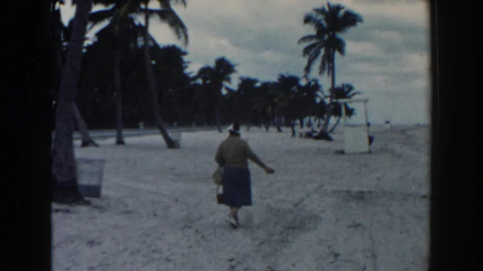 The woman walks along the Miami beach carrying a basket, with palms and a small white beach structure visible ahead.