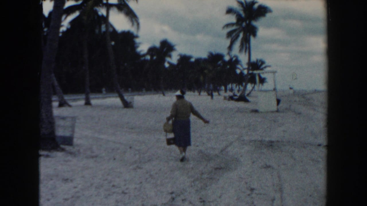 A distant home-movie view shows the woman moving across the sandy Miami beach with palms and cloudy sky behind her.