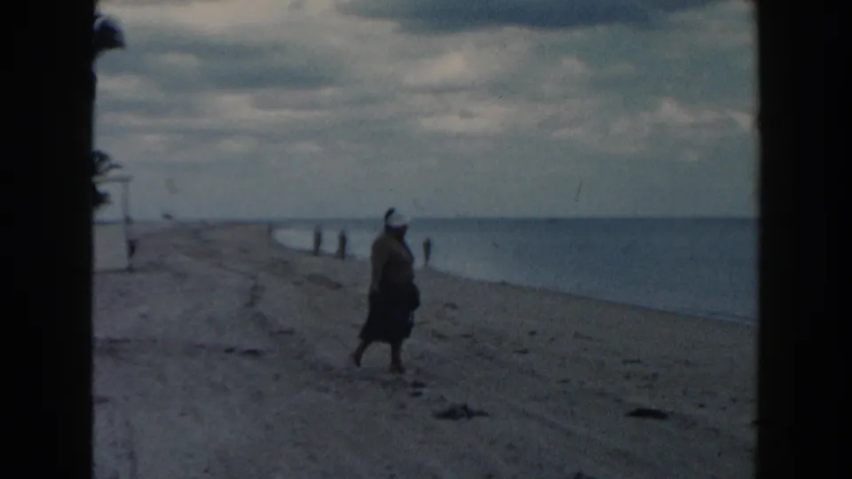 1960 Miami beach frame showing a woman in a dark skirt walking along the shoreline under a cloudy sky.