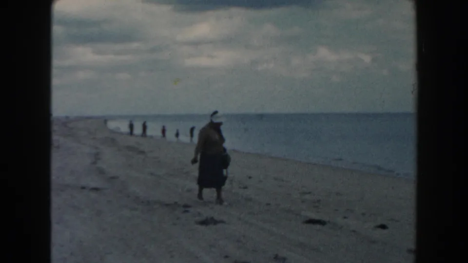 1960 Miami beach view with a woman facing the sea, distant people at the waterline and a broad cloudy horizon.