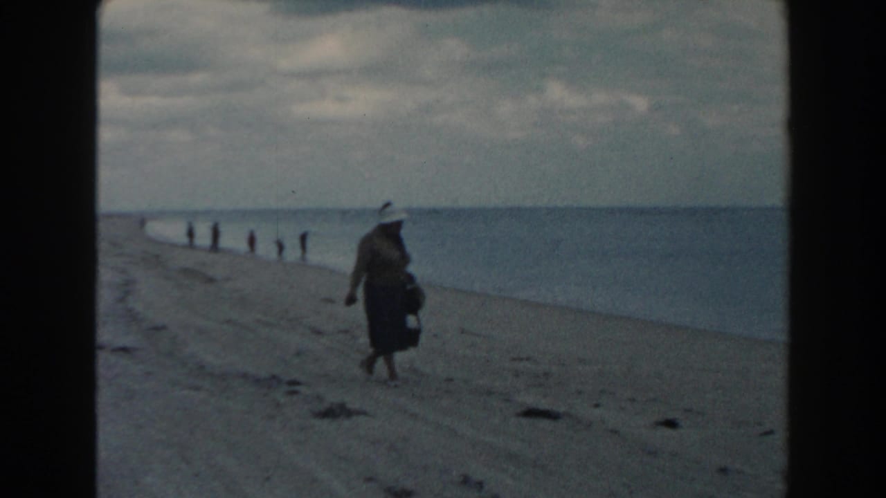 1960 Miami home-movie frame showing a woman in a light hat and long dress walking on sand beside calm ocean water.