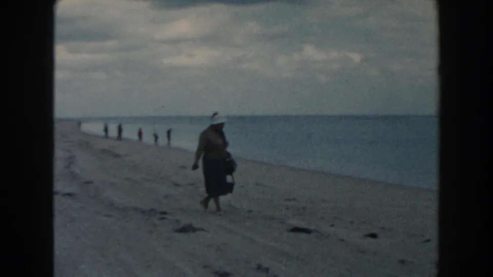 1960 Miami home-movie frame showing a woman in a light hat and long dress walking on sand beside calm ocean water.