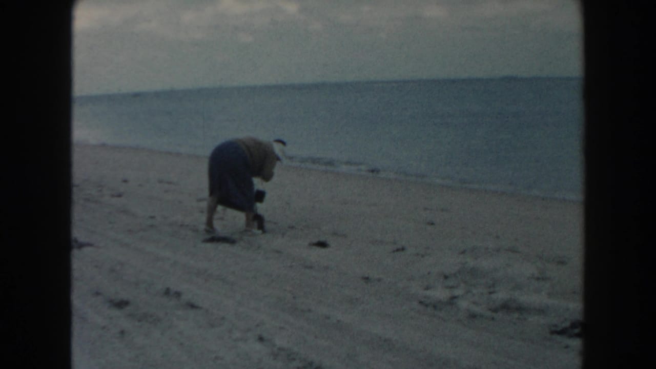 Woman bends toward the sand on a beach beside the ocean.