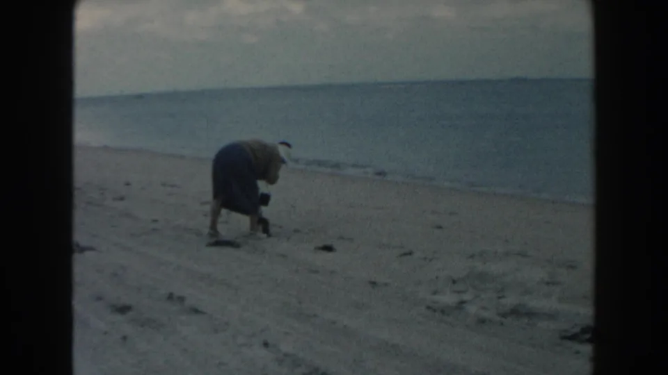 1960 Miami beach frame showing the woman leaning down near scattered sand patches with the ocean behind her.
