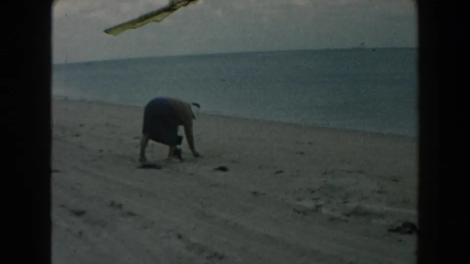 1960 Miami home-movie frame with a woman stooping low on the sand, ocean and cloudy sky behind her.