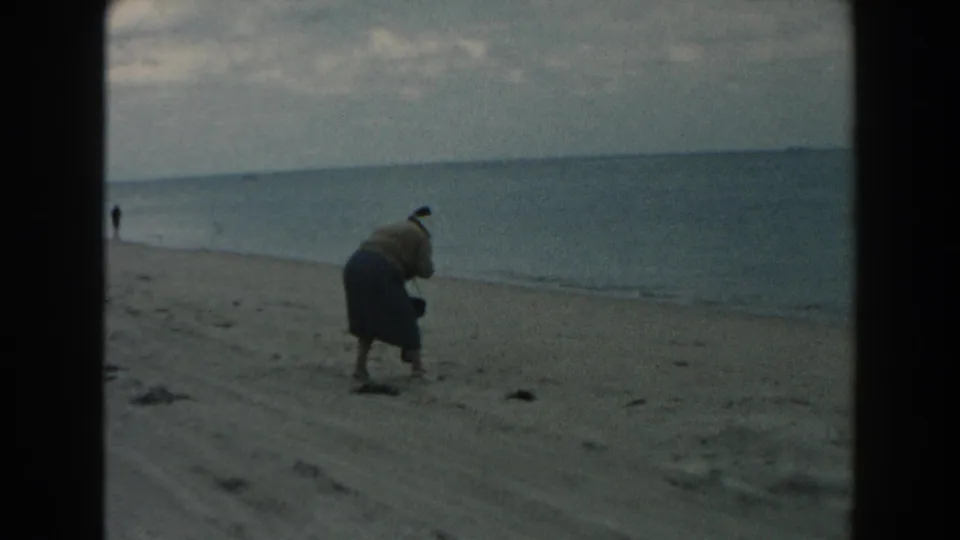 1960 Miami beach frame showing the woman bending near the sand, with calm water and a distant person along the shore.