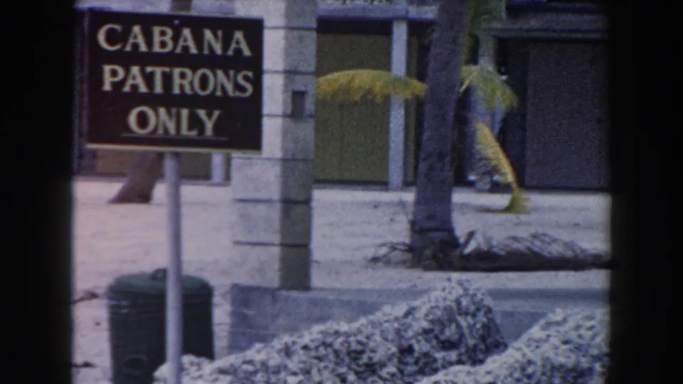 The CABANA PATRONS ONLY sign remains in view beside a palm tree, sandy ground, and cabana-like resort buildings in Miami.