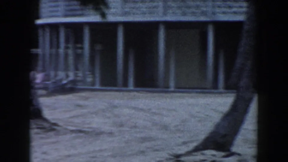 1960 Miami resort cabana area with palm trunks, sand, columns, and shaded building windows in soft 8mm color.