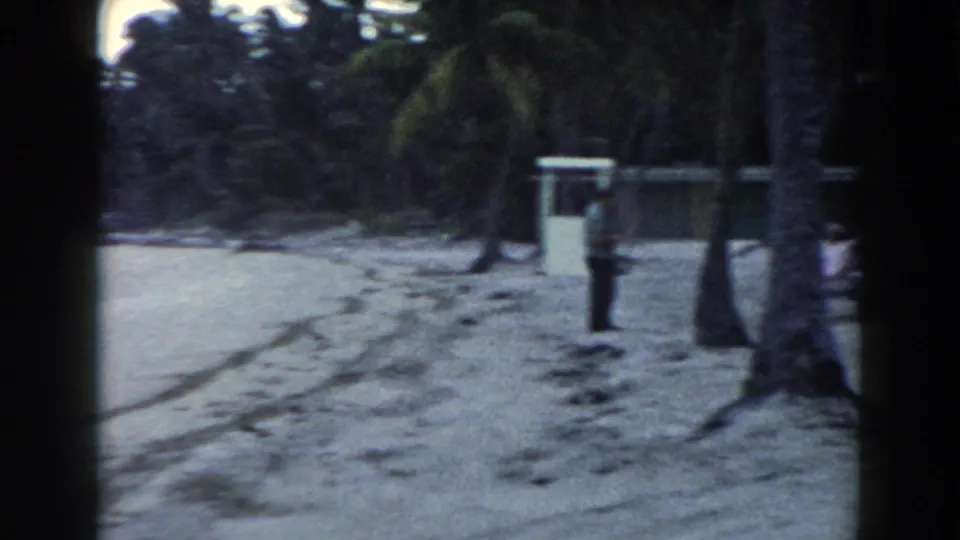 1960 Miami beachside resort area showing a sandy path, palms, a small structure, trash can, and a distant person.
