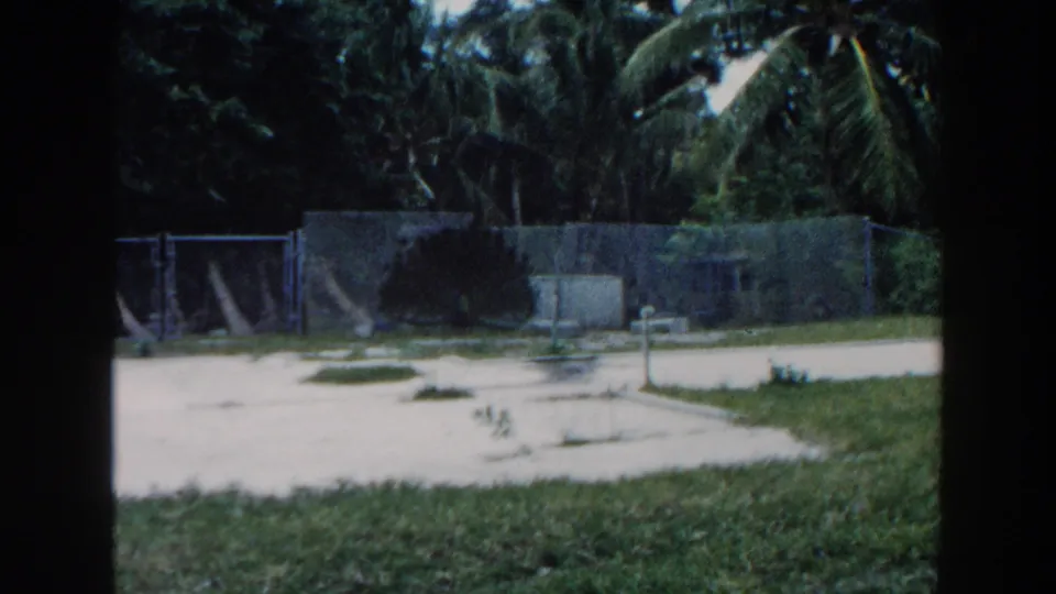 1960 Miami frame of cleared sandy ground with palm trees, fencing, posts, grass patches, and concrete foundation areas.