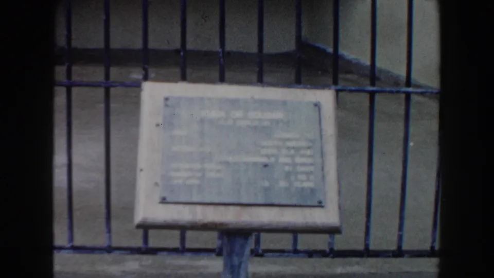 1960 Miami zoo home-movie frame focusing on a pale information sign inside or in front of a barred animal enclosure.