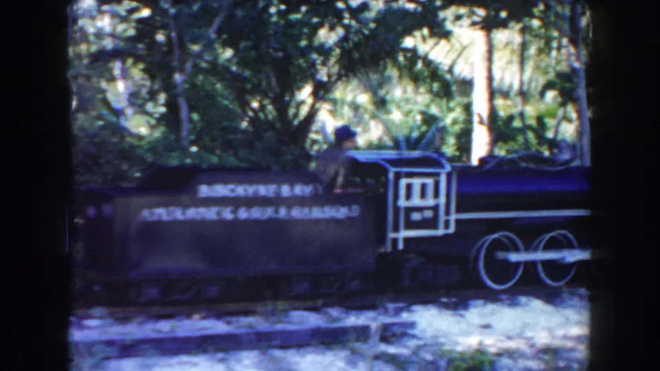 A miniature train passenger car with readable lettering passes through a leafy Miami park or zoo area in the 1960 home-movie clip.