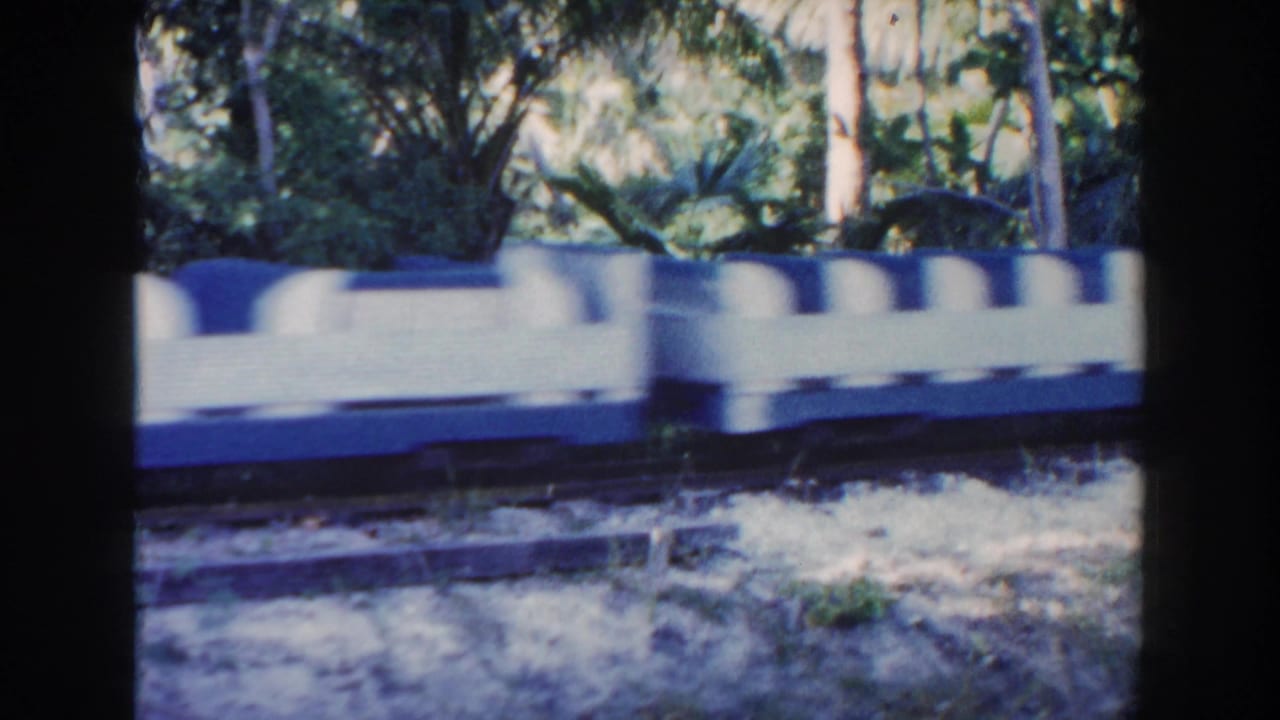 1960 Miami home-movie frame of a blue and white amusement park train car moving along tracks through a wooded park setting.