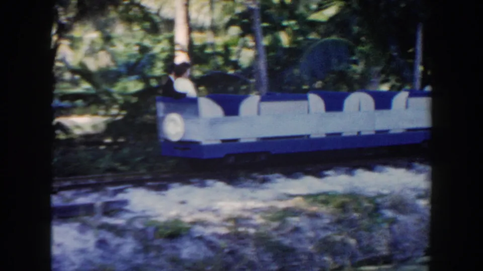 Open amusement park train car in blue and white passes through a leafy Miami setting, with a seated rider visible at a distance.