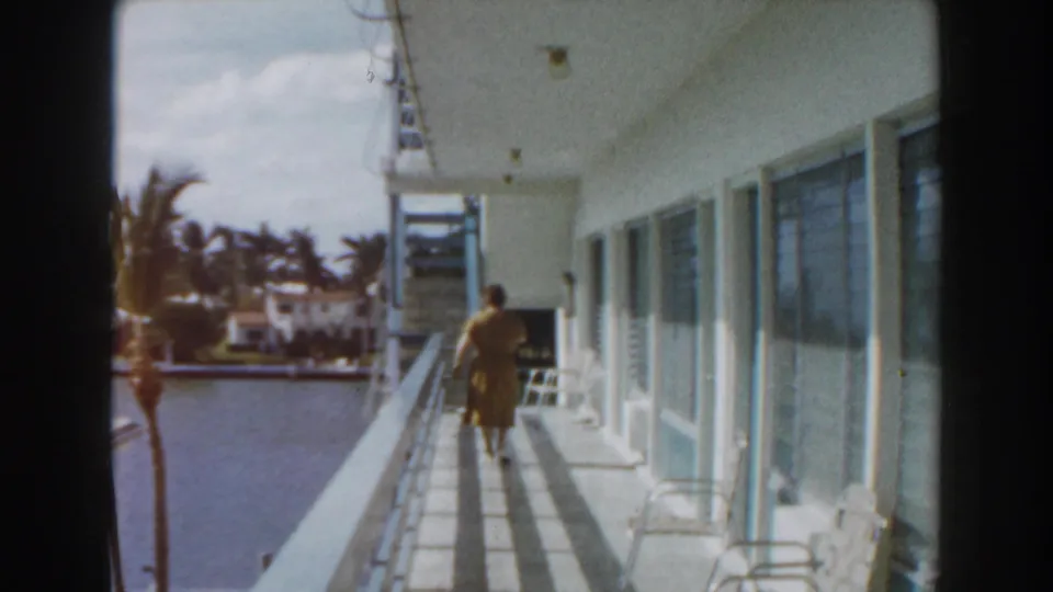 Woman in a yellow dress walks along a Miami waterfront balcony, with canal water, palms, and houses in the background.